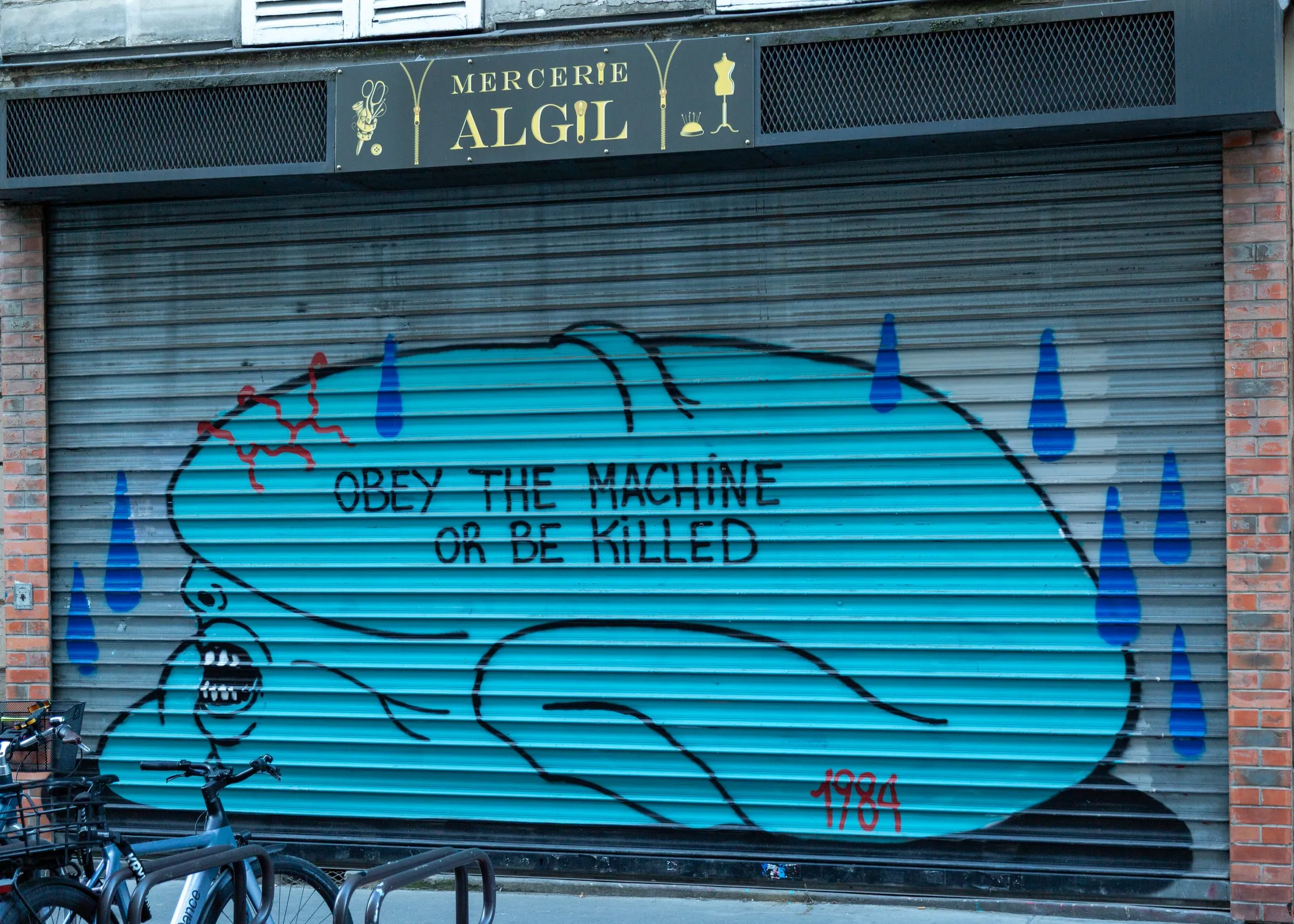 "Obey the machine or be killed": a painted shutter below a mercerie sign